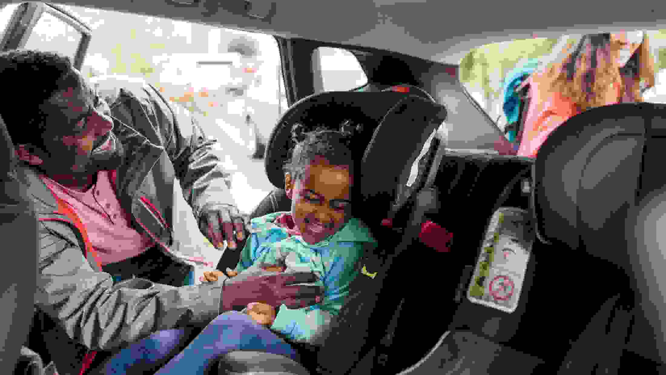Black male father buckles his young daughter into a car seat while tickling her ahead of a road trip