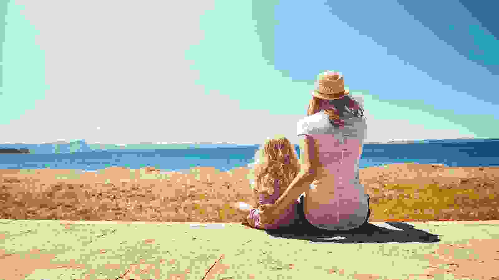 Mother and daughter sat on the beach on a sunny day looking out to sea