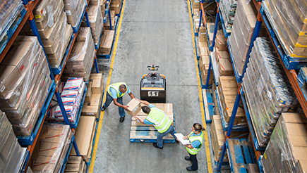 Three people in High Vis Vests in a warehouse, starting to stack boxes on a wooden pallet.