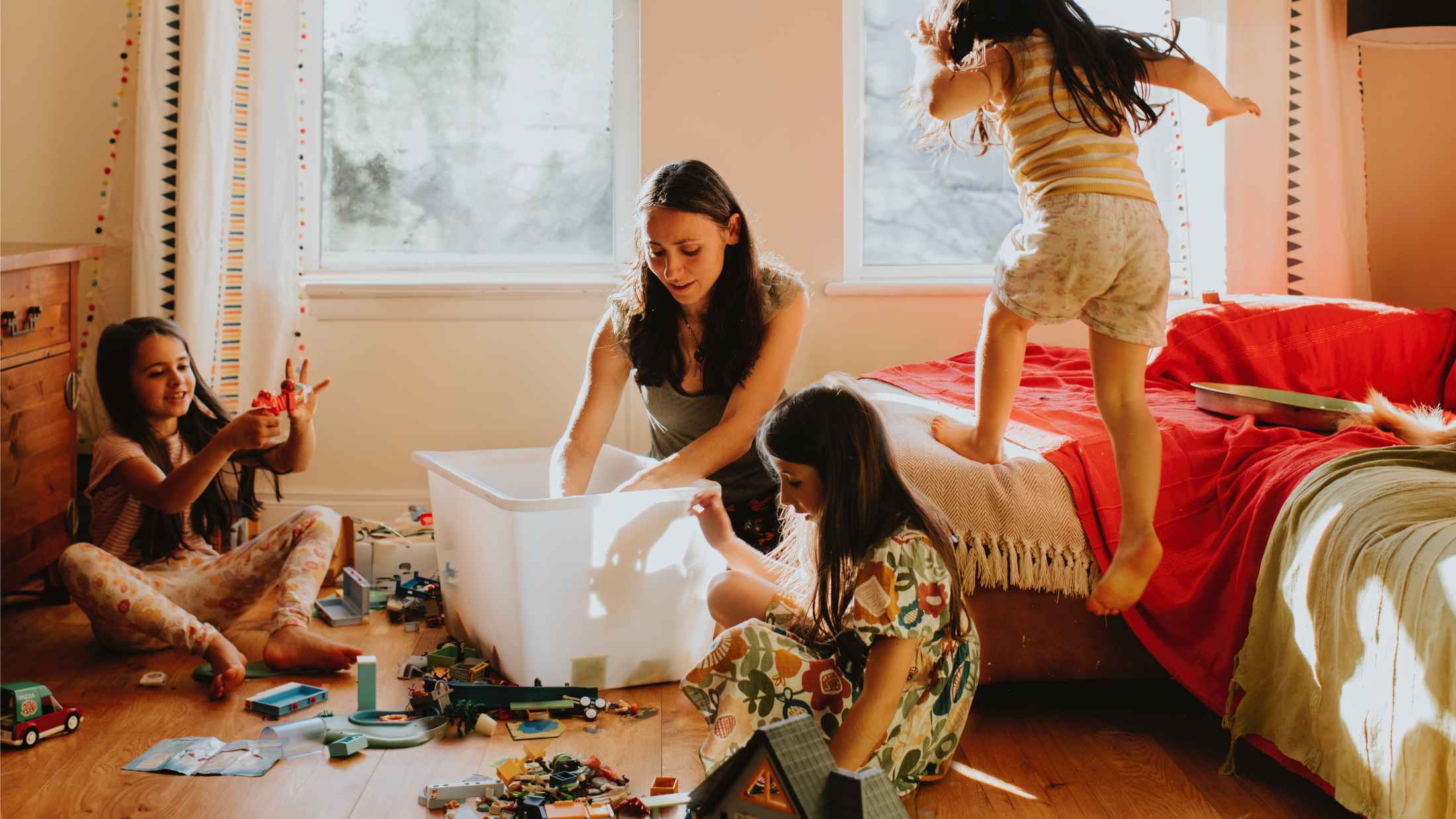 A loving mother sits on the floor in a sunny room with her three young daughters rummaging through a box of toys
