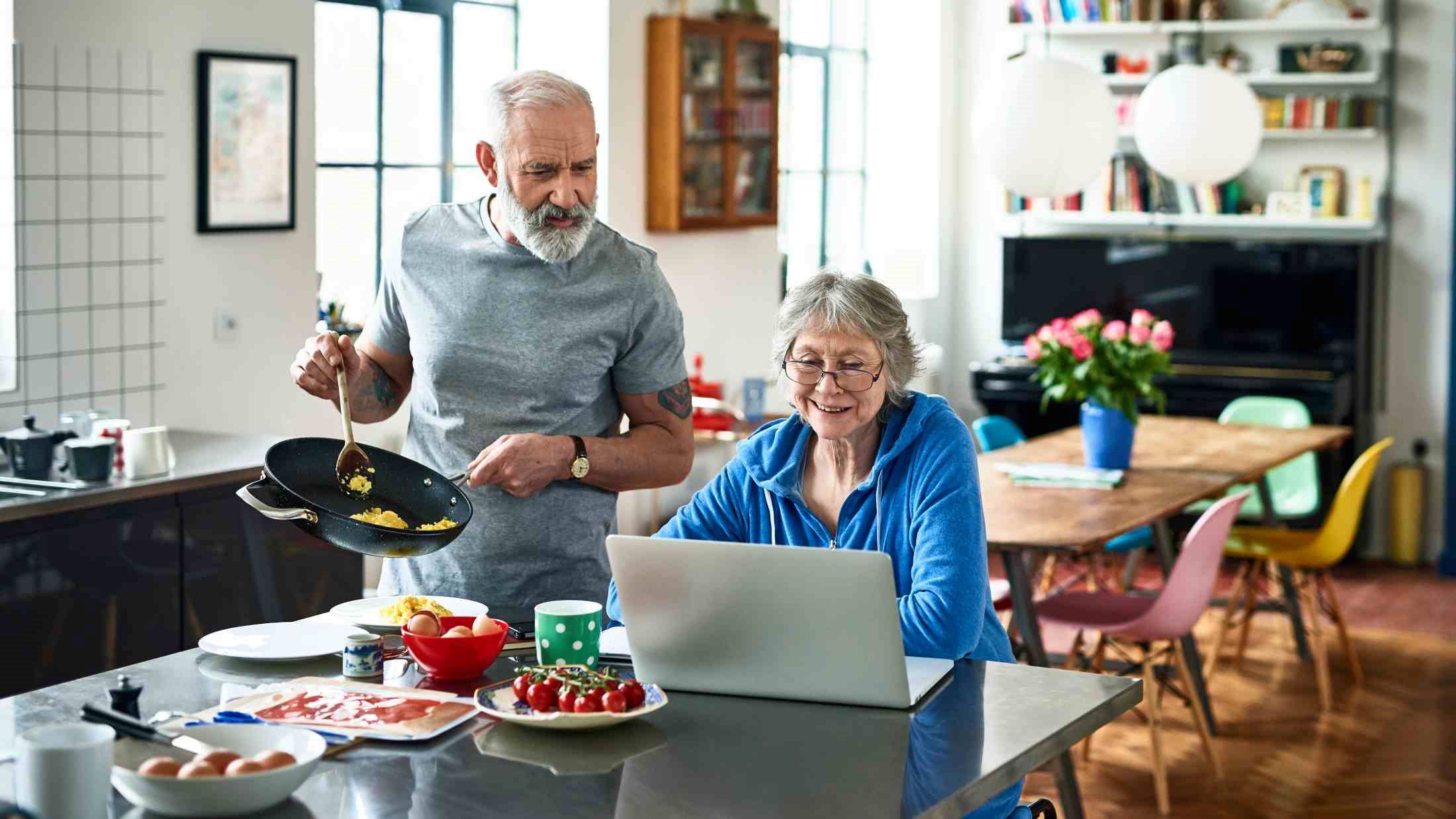 Retired couple in modern kitchen on the weekend, woman surfing the web, man holding frying pan and serving scrambled eggs