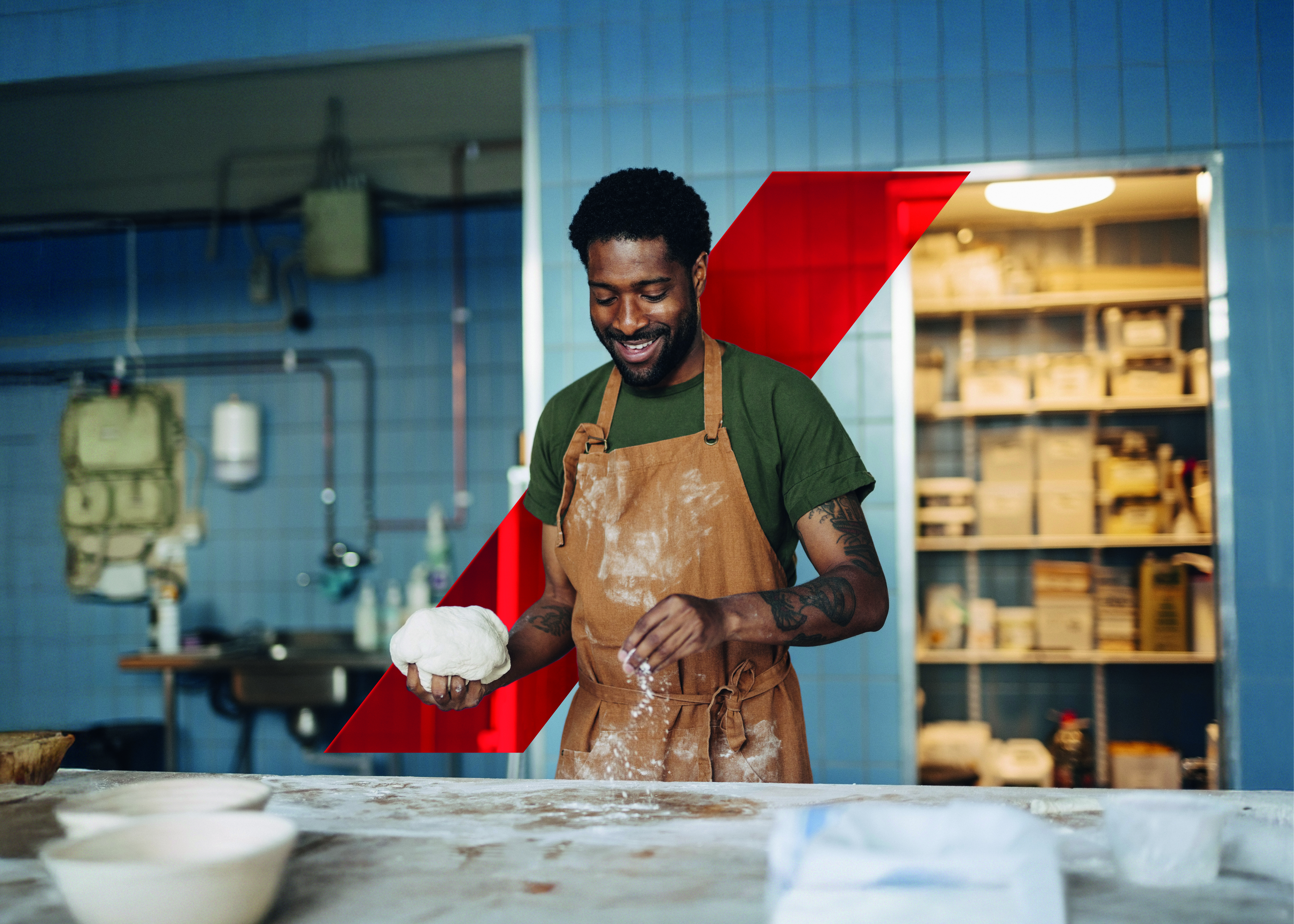Smiling male baker dusting flour on table and holding dough while working at bakery