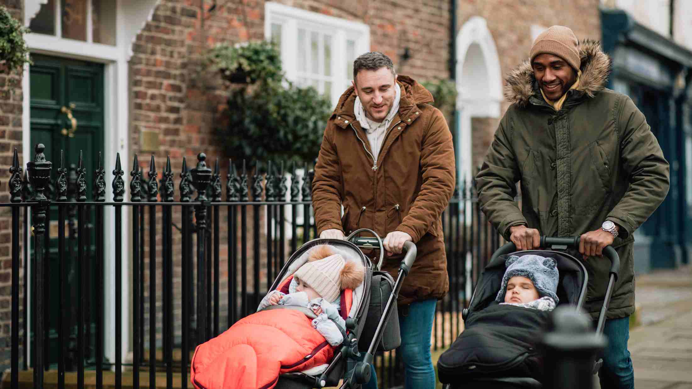 Two male friends are out in Tynemouth, North East UK walking on a pavement and pushing their baby sons in strollers