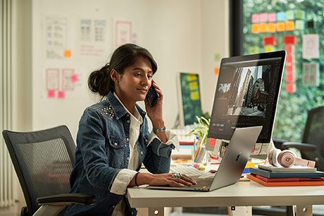 Woman at desk holding phone while using laptop