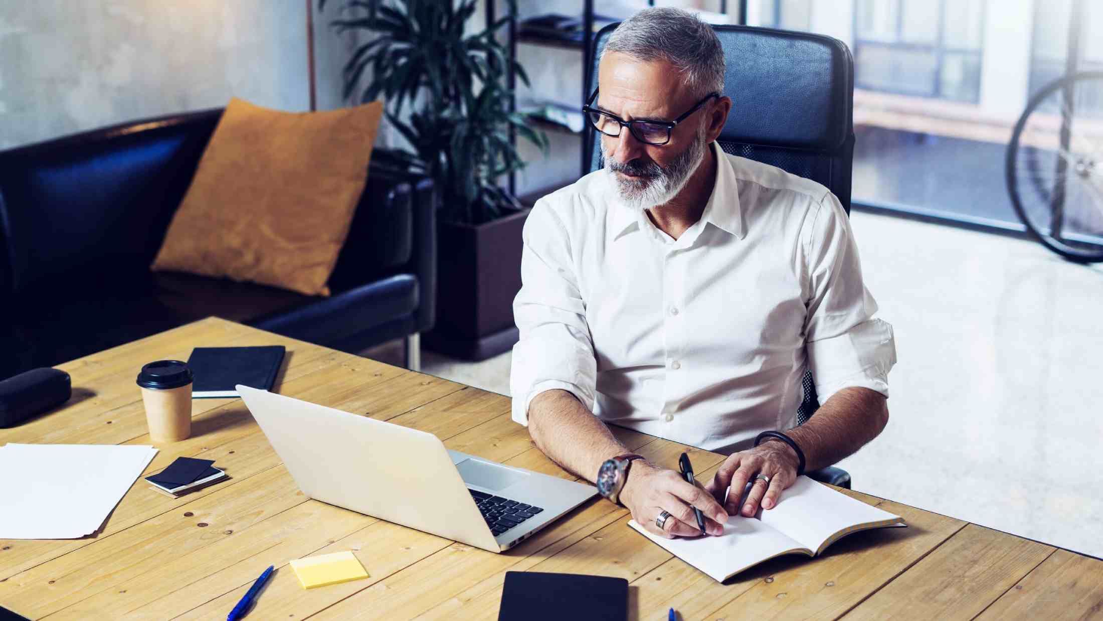 Man with greying hair working on his laptop while making notes