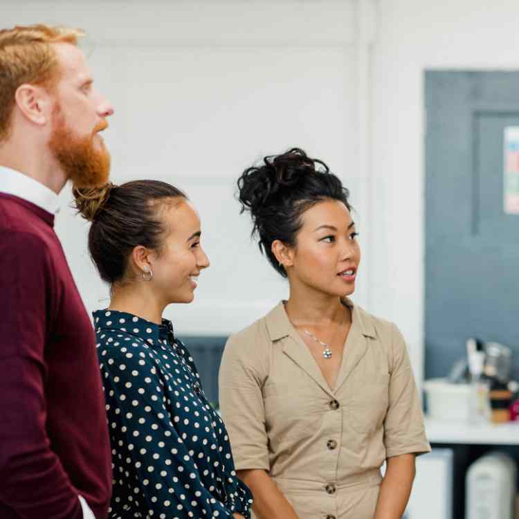 Colleagues standing in a small group discussing something while one woman gestures with her hands holding a white board pen