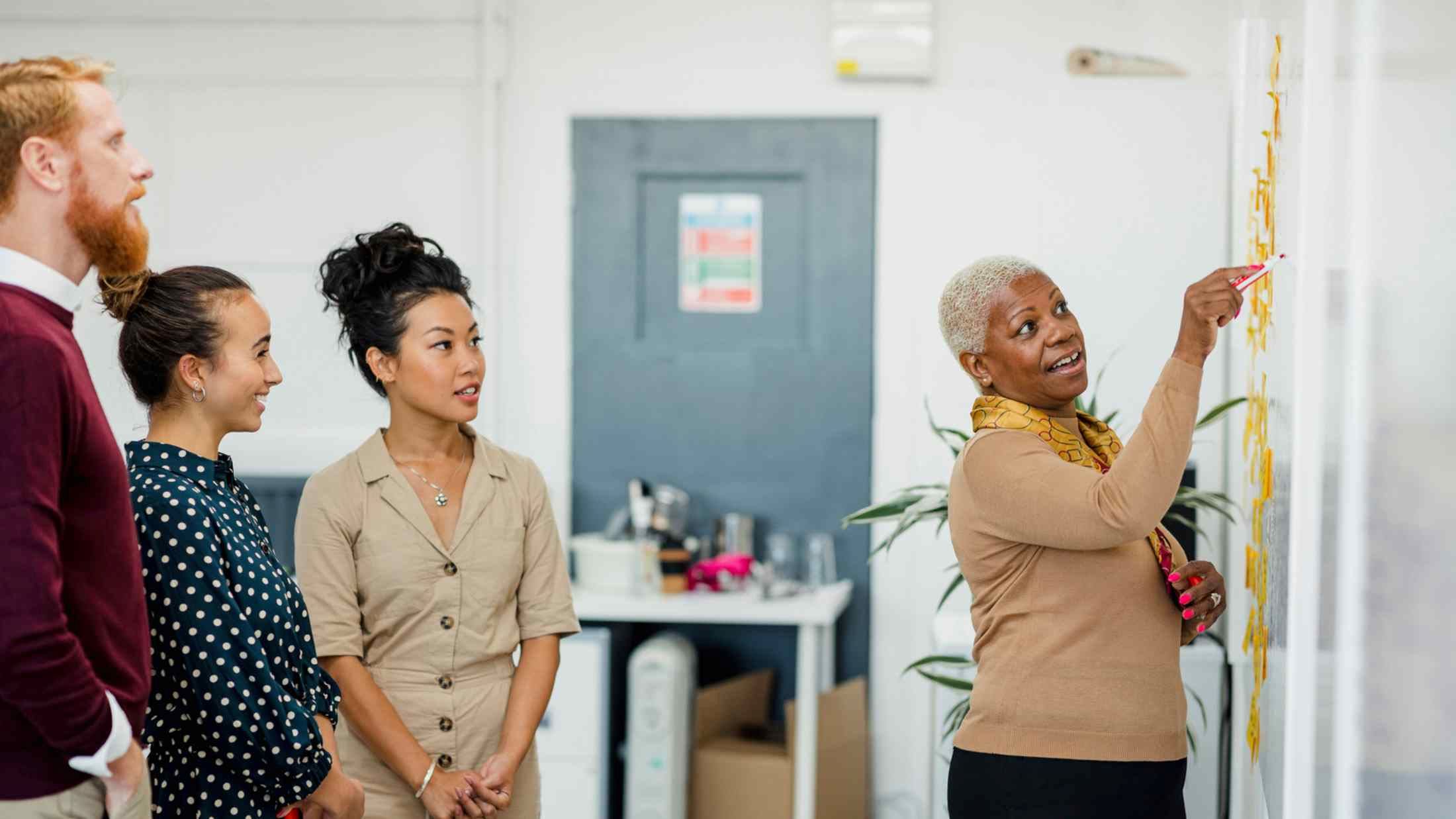 Colleagues standing in a small group discussing something while one women gestures with her hands holding a white board pen
