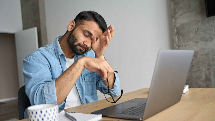 Exhausted business man rubbing eyes sitting in modern home office with laptop on desk