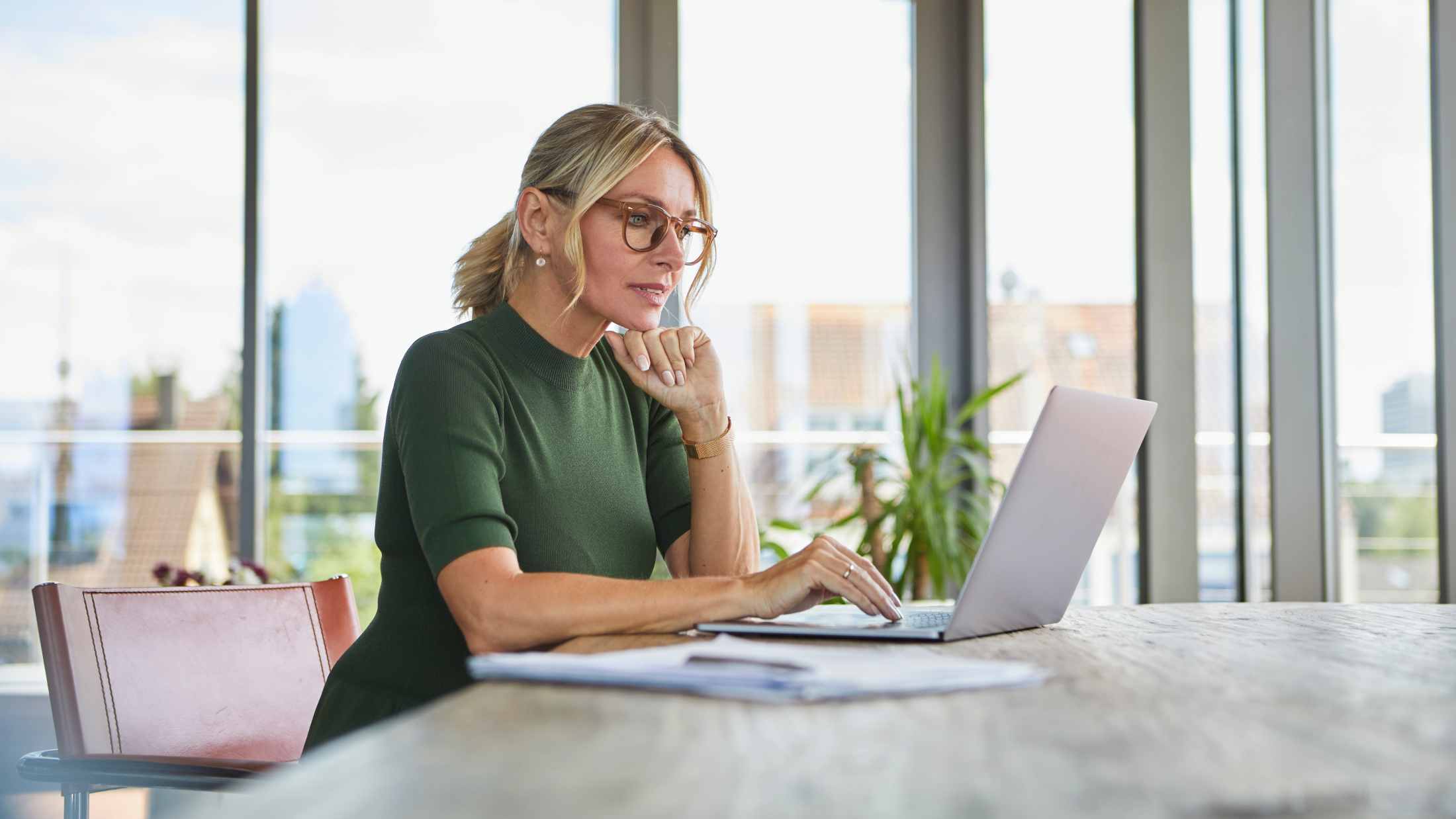 A professional blonde woman wearing glasses and a green top sits at a wooden desk while focused on working on her laptop in a bright, modern office.