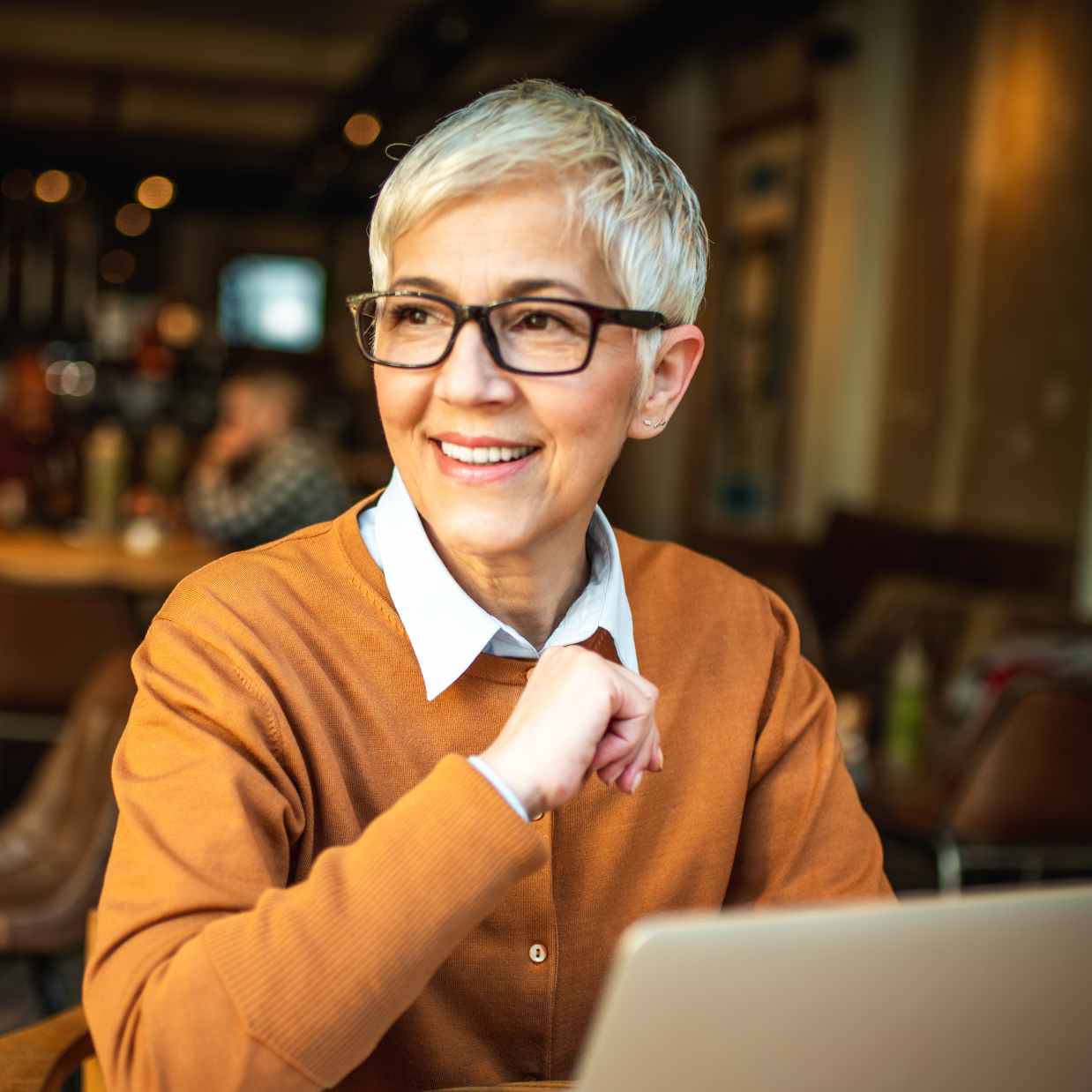 Middle aged woman with short hair and glasses wearing an orange jumper using her laptop while in a coffee shop