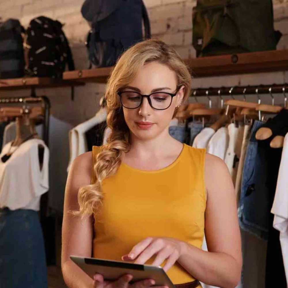 Womena wearning a mustard coloured sleeveless top using an iPad while stood in her clothes shop
