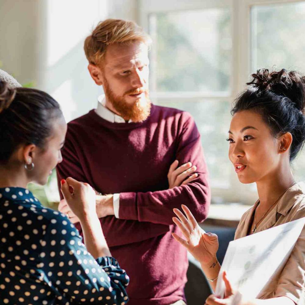 Colleagues standing in a small group discussing something. One of the women is holding documents and gesturing with her hands as the others watch and listen.