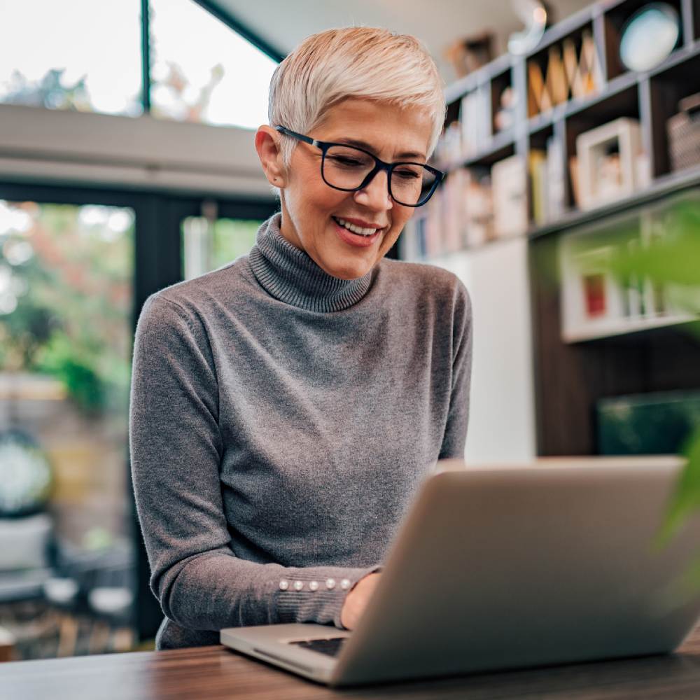 Modern mature woman with glasses wearing grey turtle neck top using laptop at home