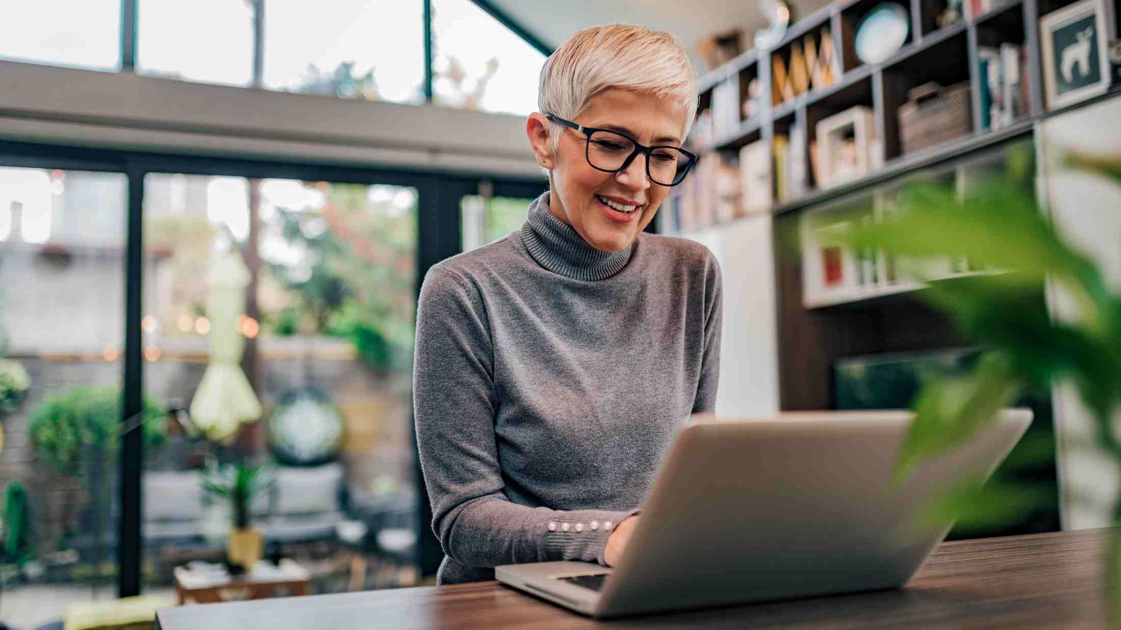 Modern mature woman with glasses wearing grey turtle neck top using laptop at home