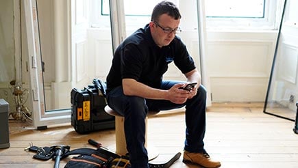 Ian, late 30's male, sits while using his phone. He is surrounded by tools in a home that is bare. 