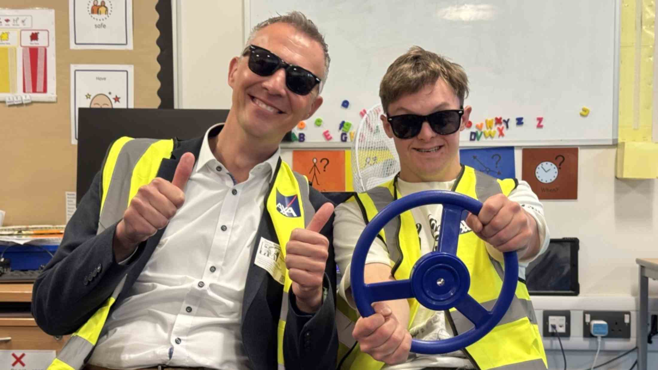 Two men in a classroom, wearing safety vests and sunglasses, engaging in a role-playing activity with a steering wheel, surrounded by educational materials.