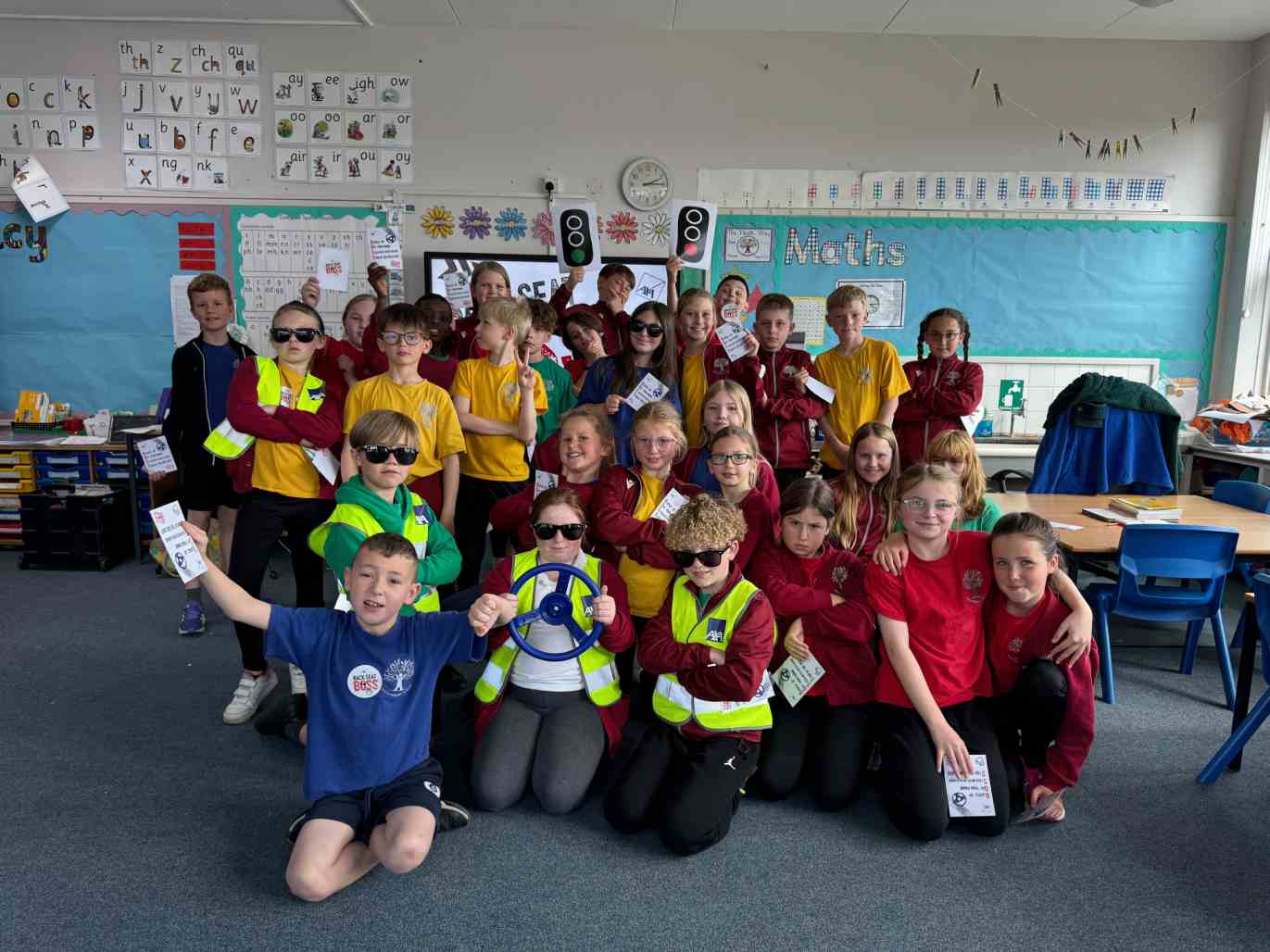 KS2 students in a road safety lesson, wearing bright vests and holding a steering wheel props and cards, in a classroom with a traffic signal and educational displays