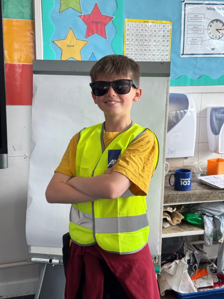 A young boy student stands in front of a whiteboard, wearing sunglasses, a bright yellow safety vest, and a maroon jacket, with his arms crossed.