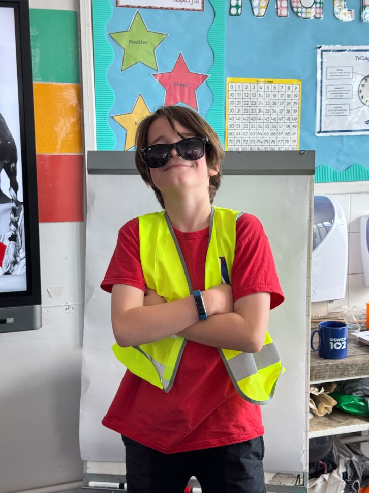 A boy student stands in a classroom, wearing sunglasses, a red t-shirt, and a bright yellow safety vest, with his arms crossed