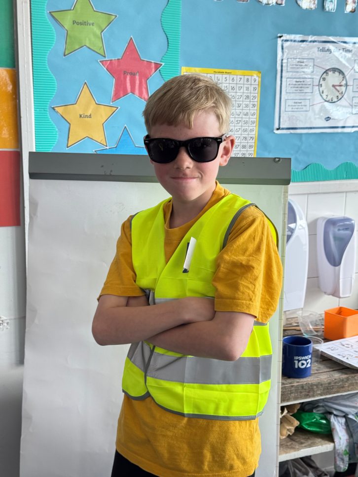 A young boy with short light brown hair wears sunglasses and a bright yellow safety vest over a mustard-yellow t-shirt, with his arms crossed