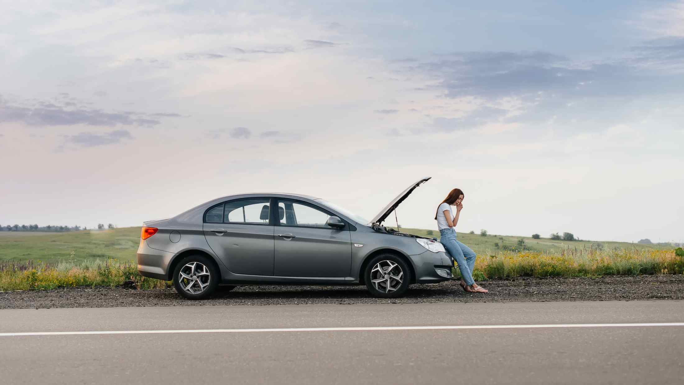 A frustrated young girl stands near a broken-down car at the side of the road