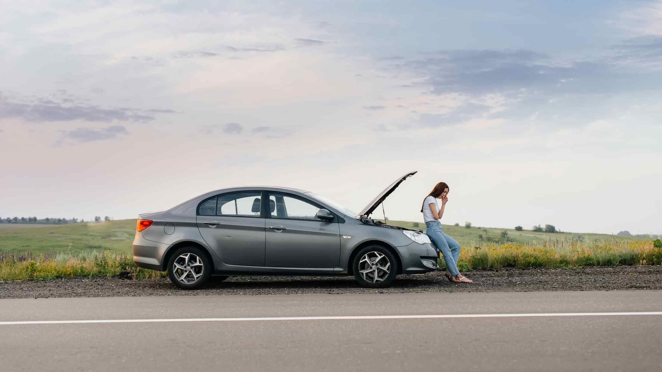 A frustrated young girl stands near a broken-down car at the side of the road