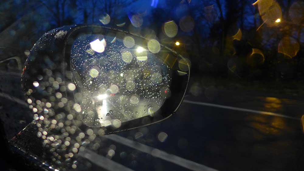 View out of the side of a car at night with water droplets visible via reflected light on the window and side mirror