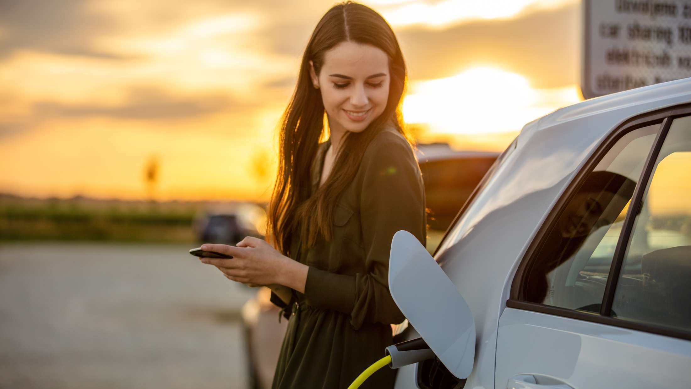 Woman waits for electric car to charge in parking lot at sunset
