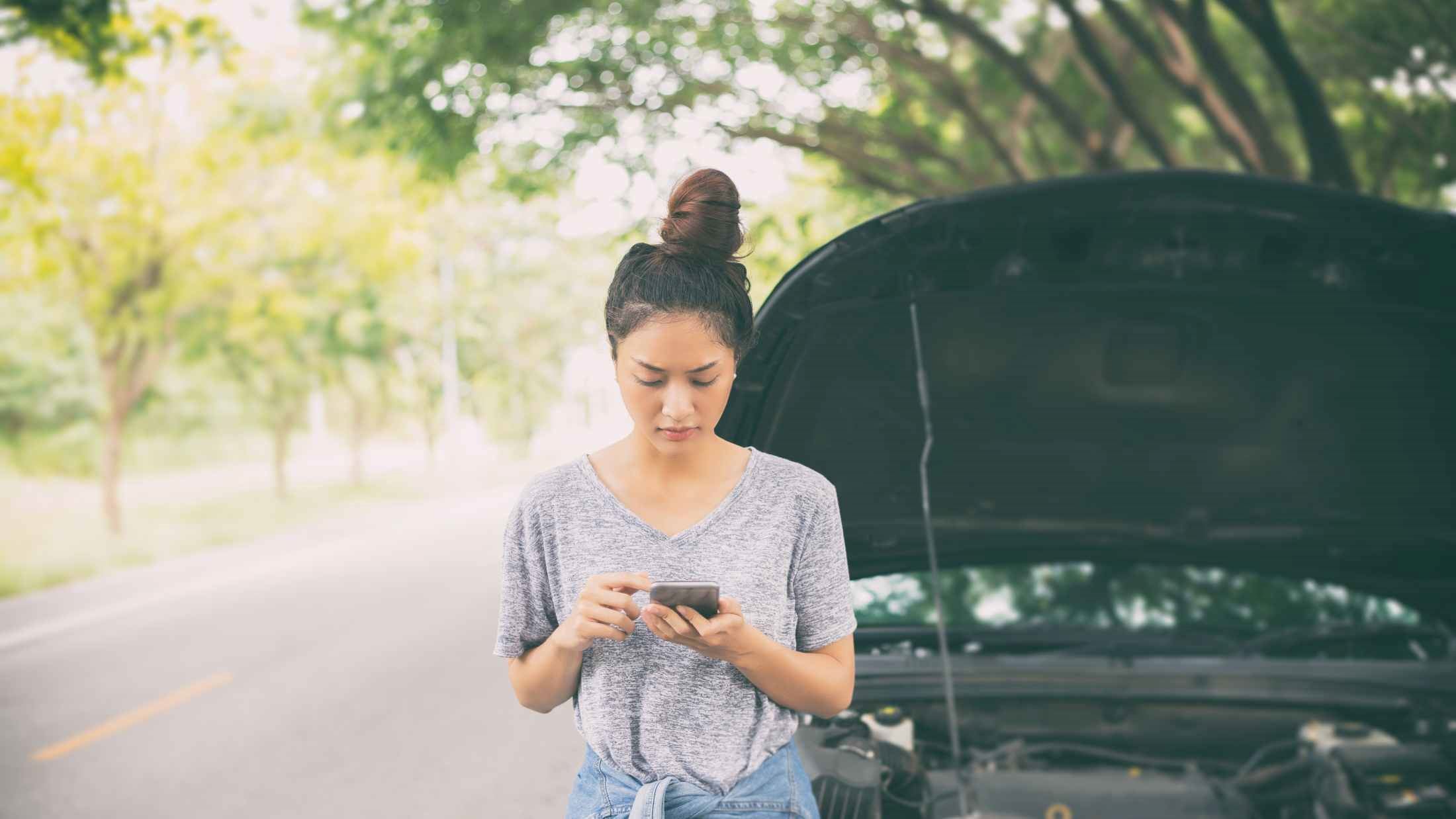 Young woman using mobile phone next to her breakdown car on the road