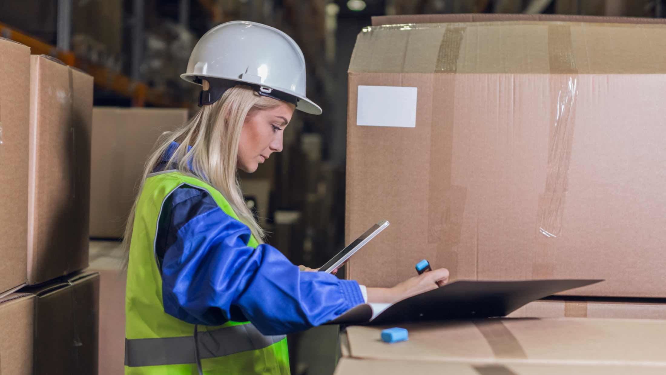 Woman wearing hi-vis jacket and hard hat holding phone and highlighter pen surrounded by cardboard boxes