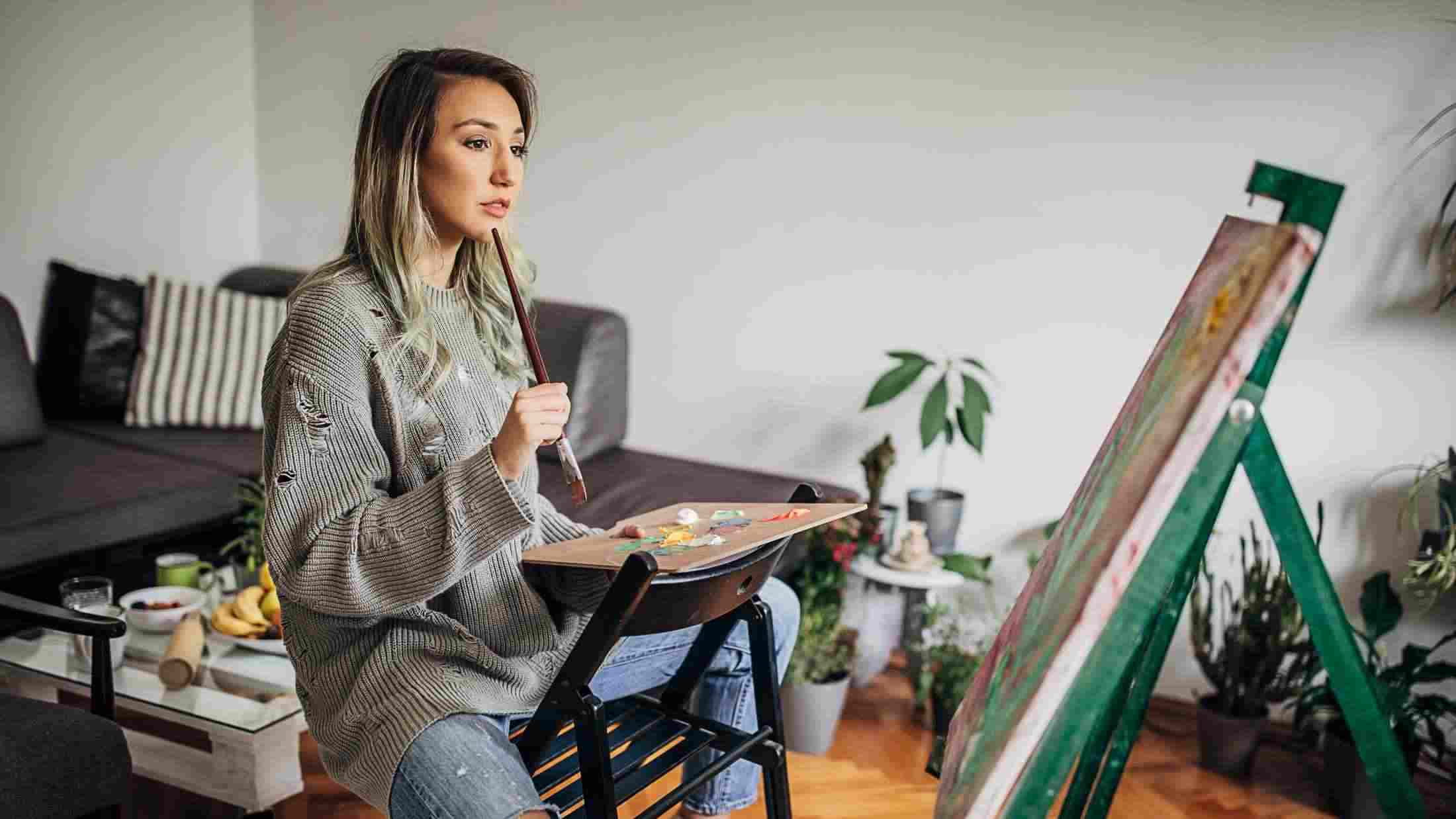 Female painter sat on a chair holding a paint brush while looking at a canvas