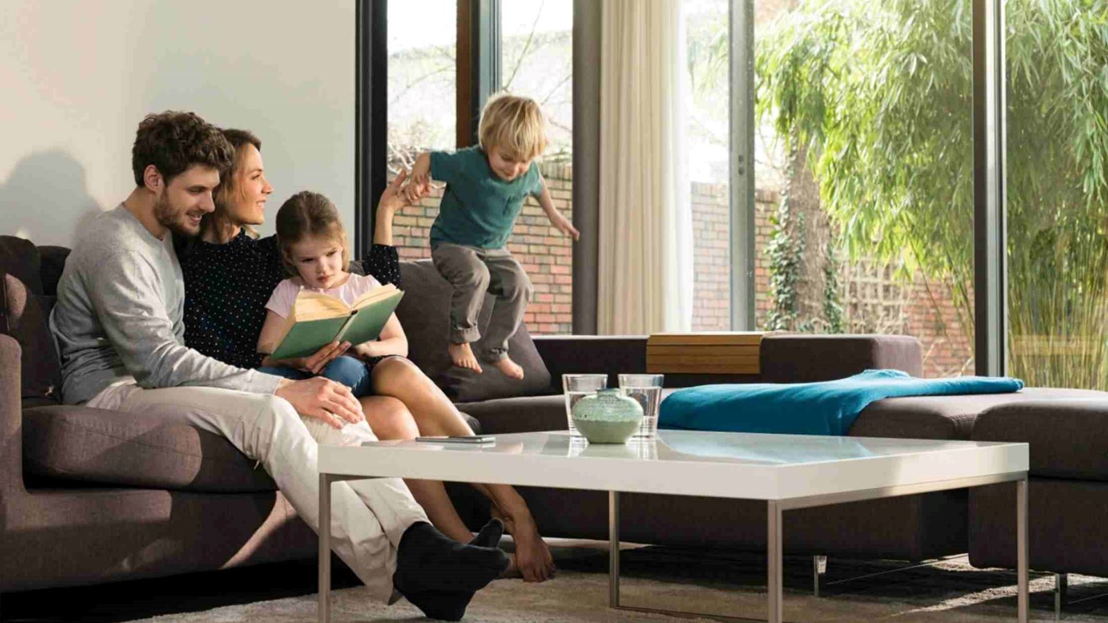 Family sat on sofa while daughter reads a book and son jumps up and down