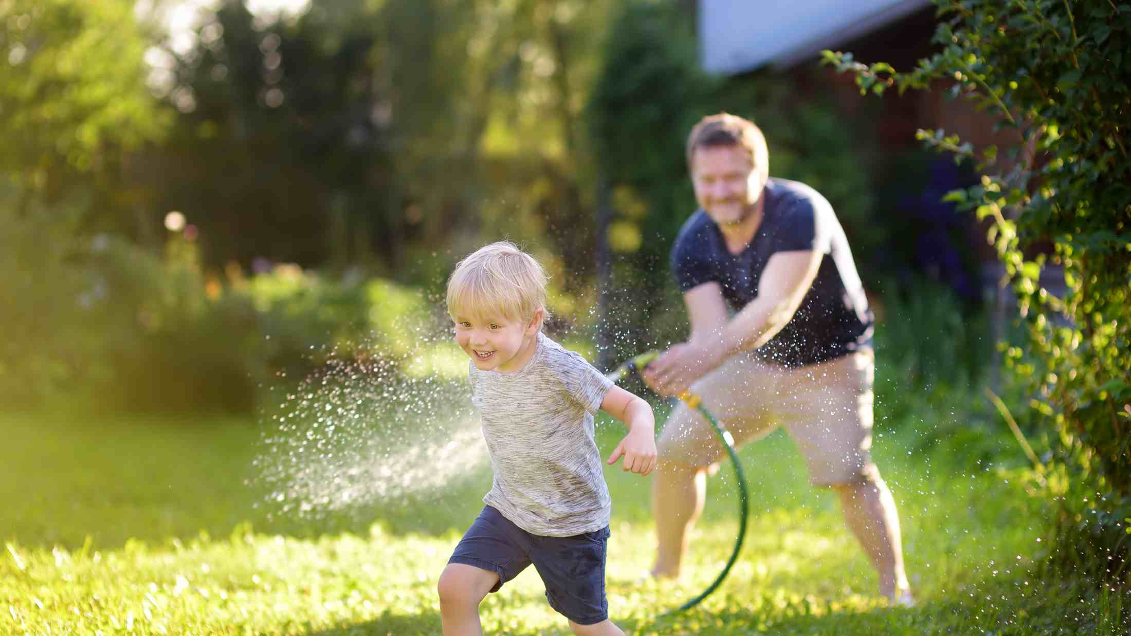 Funny little boy with his father playing with garden hose in sunny backyard