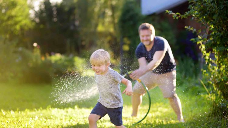 Funny little boy with his father playing with garden hose in sunny backyard