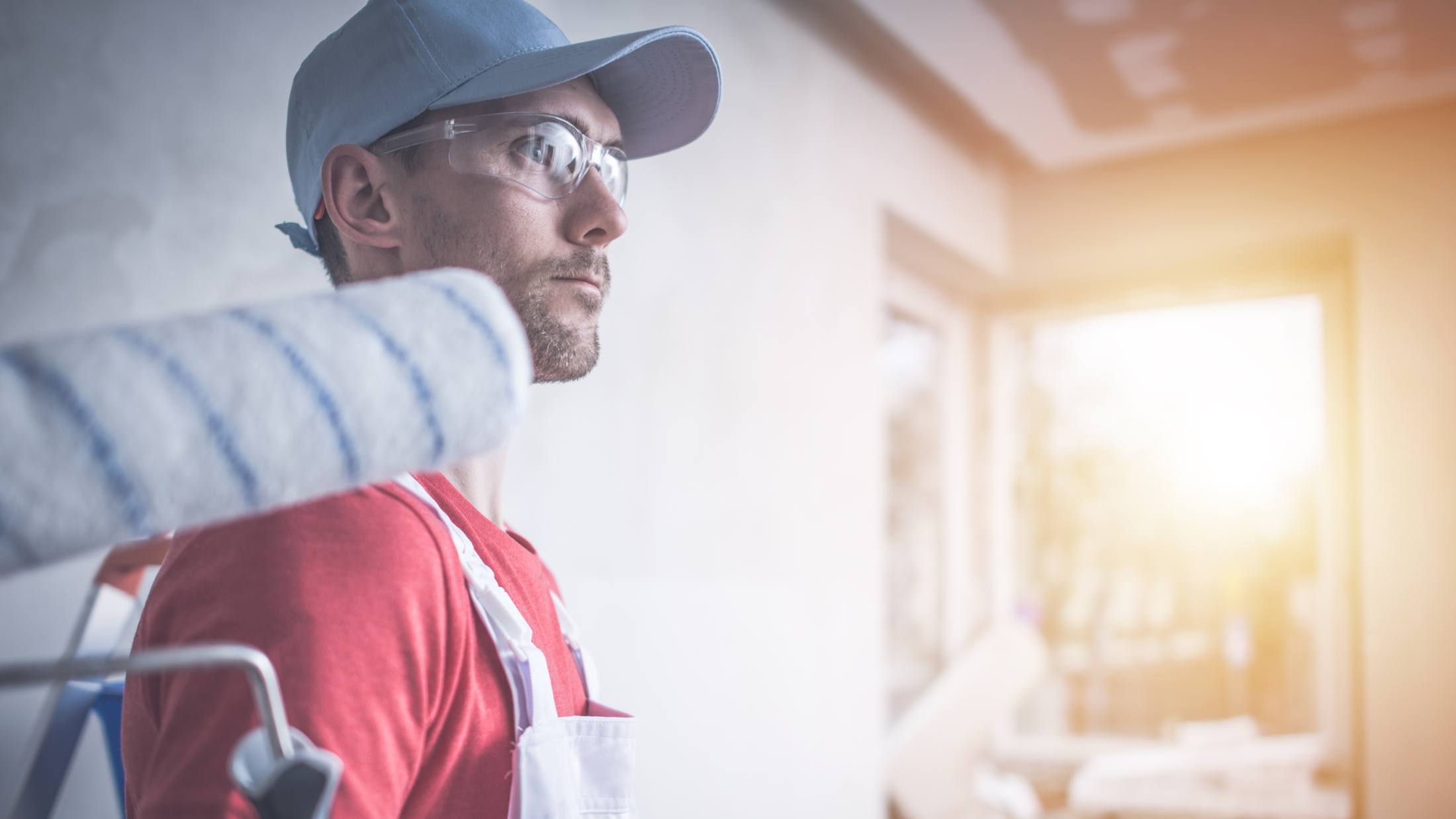 Man wearing blue cap, a red t-shirt, and white overalls, holding a roller while painting a room