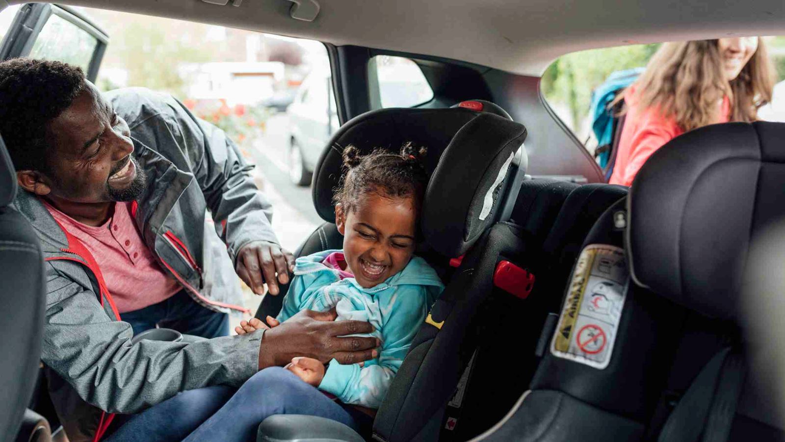 Black male father buckles his young daughter into a car seat while tickling her ahead of a road trip