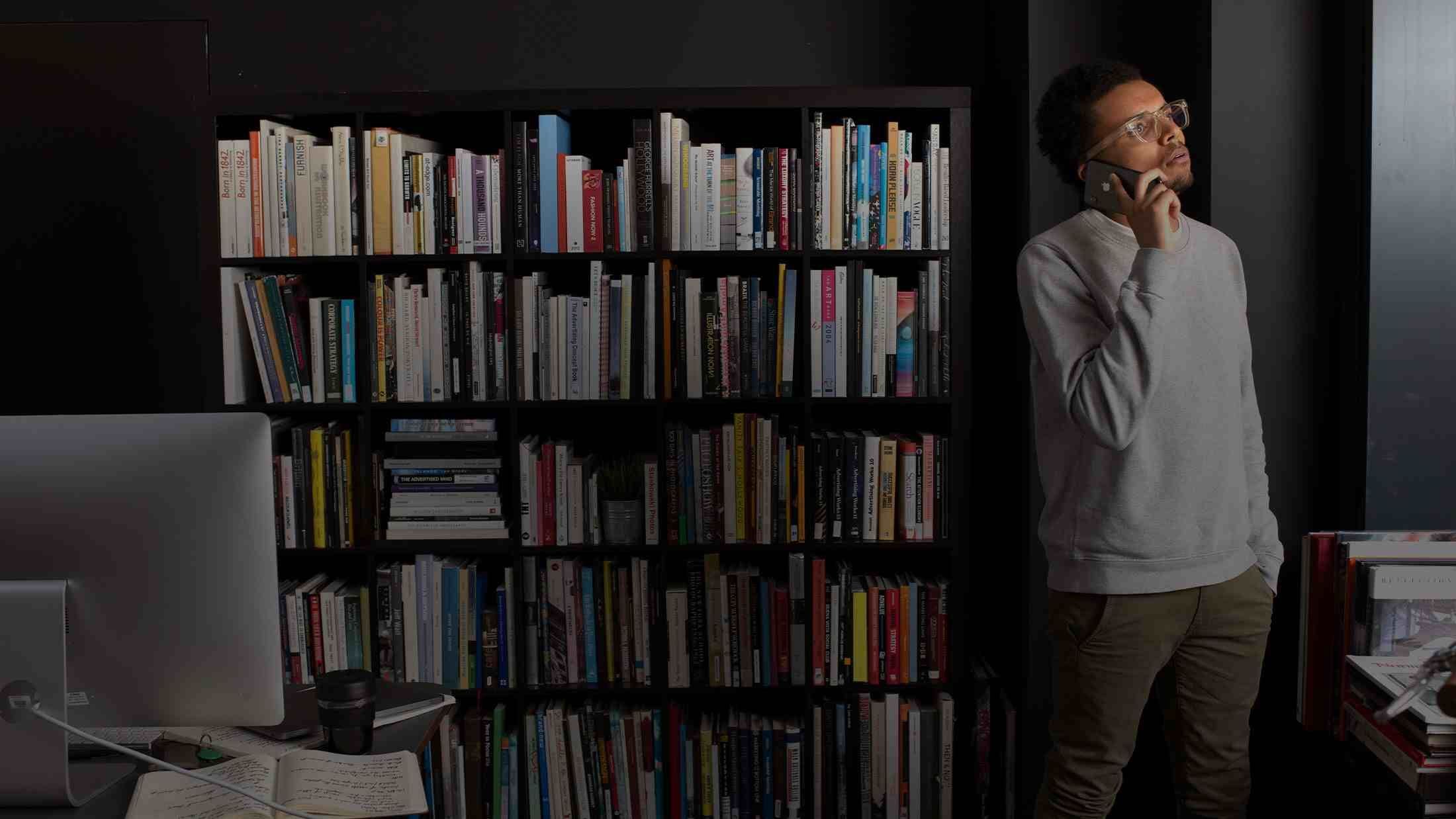 Young black man on his mobile phone while stood in his home office looking out of the window with bookshelf in background