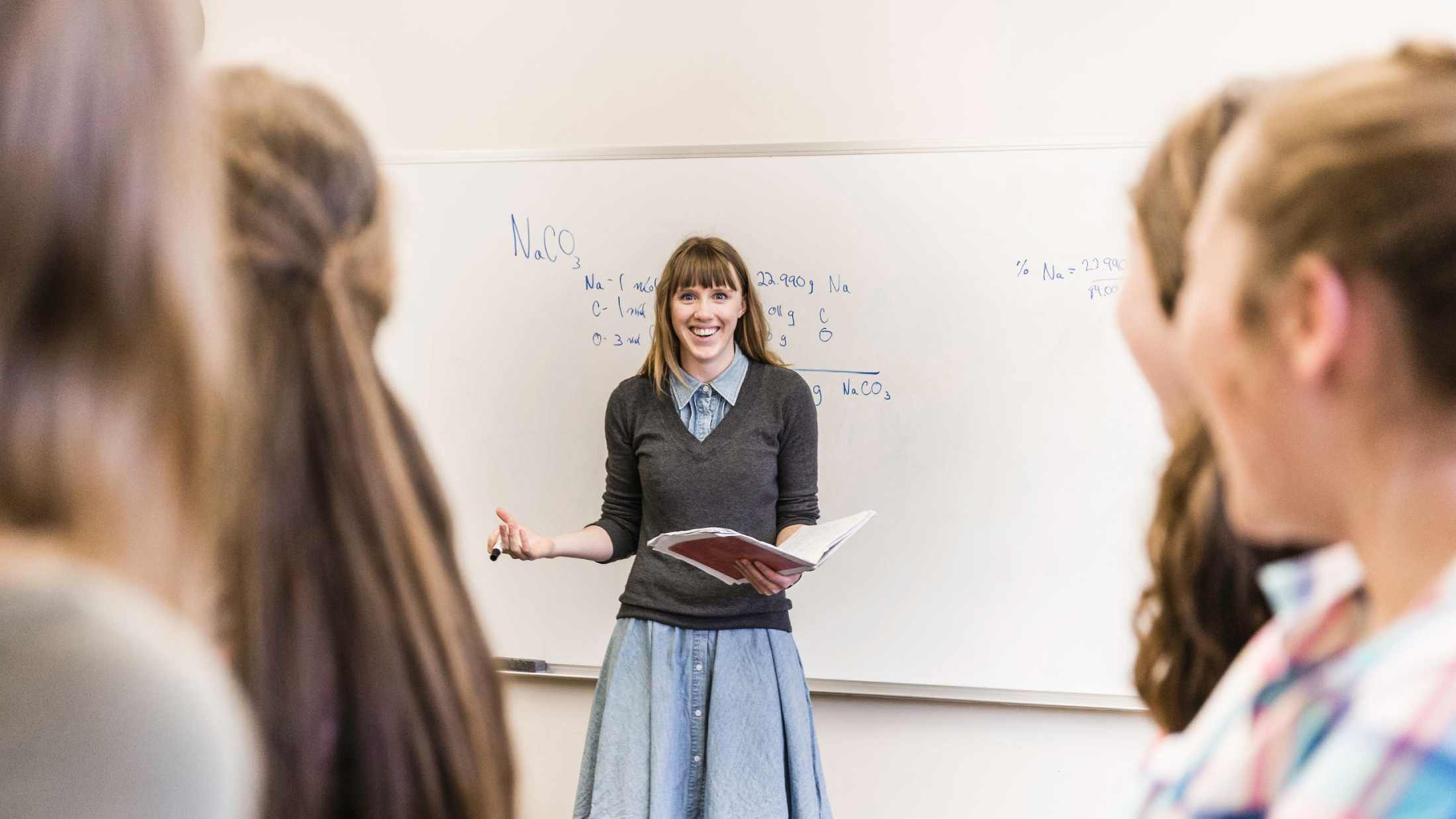 Chemistry teacher wearing blue denim dress and grey v-neck tshirt stood in front of a whiteboard