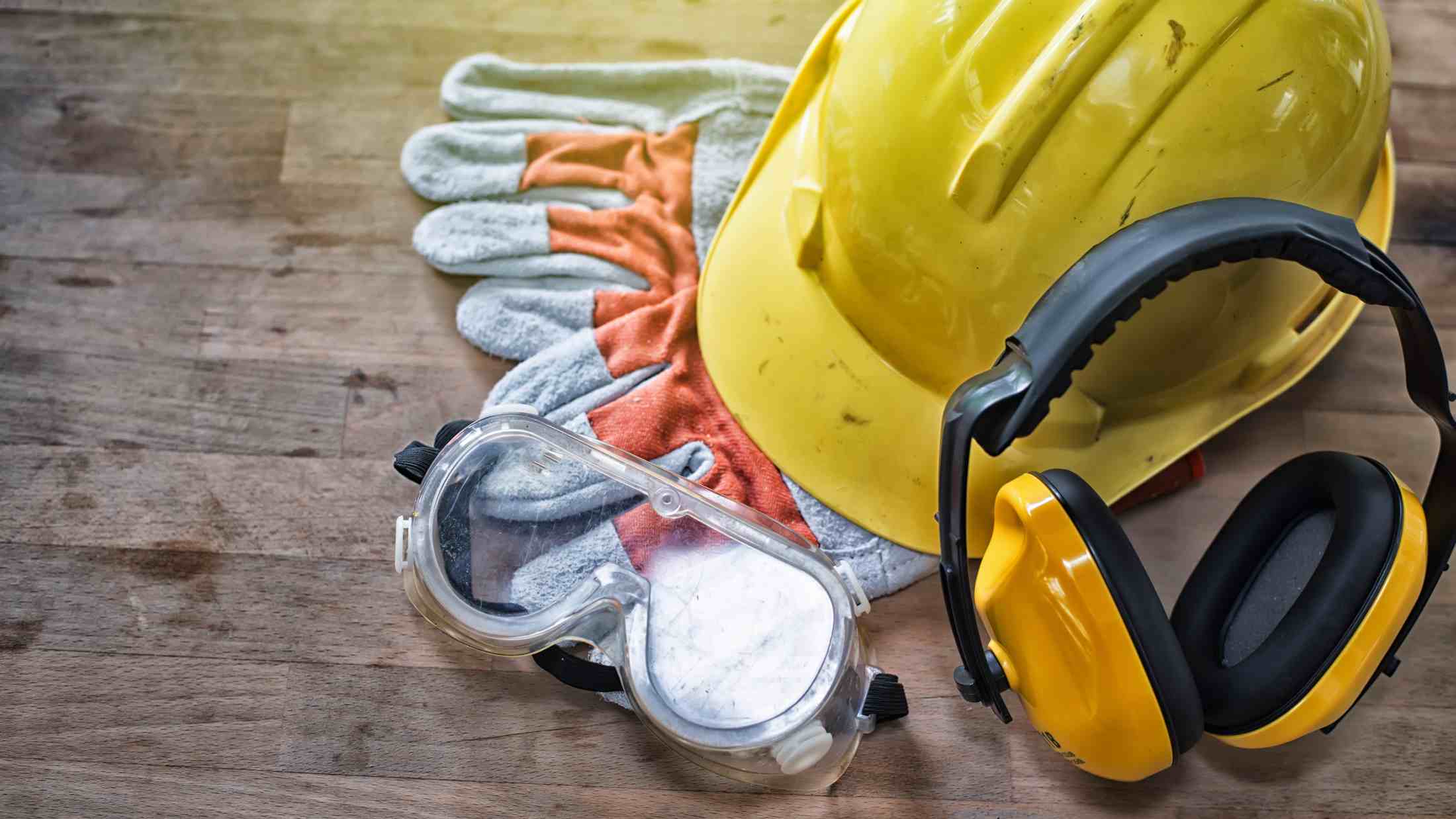 Yellow hard hat, ear defenders, leather gloves and protective goggles on wooden board