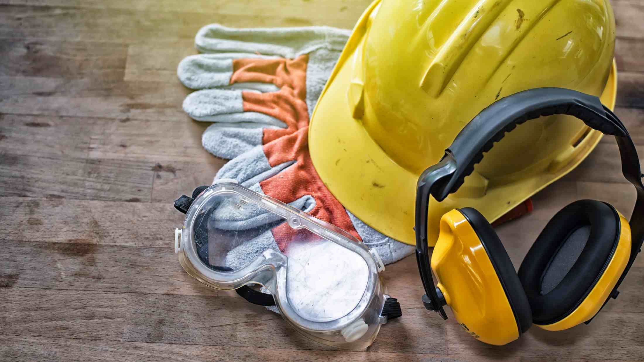 Yellow hard hat, ear defenders, leather gloves and protective goggles on wooden board