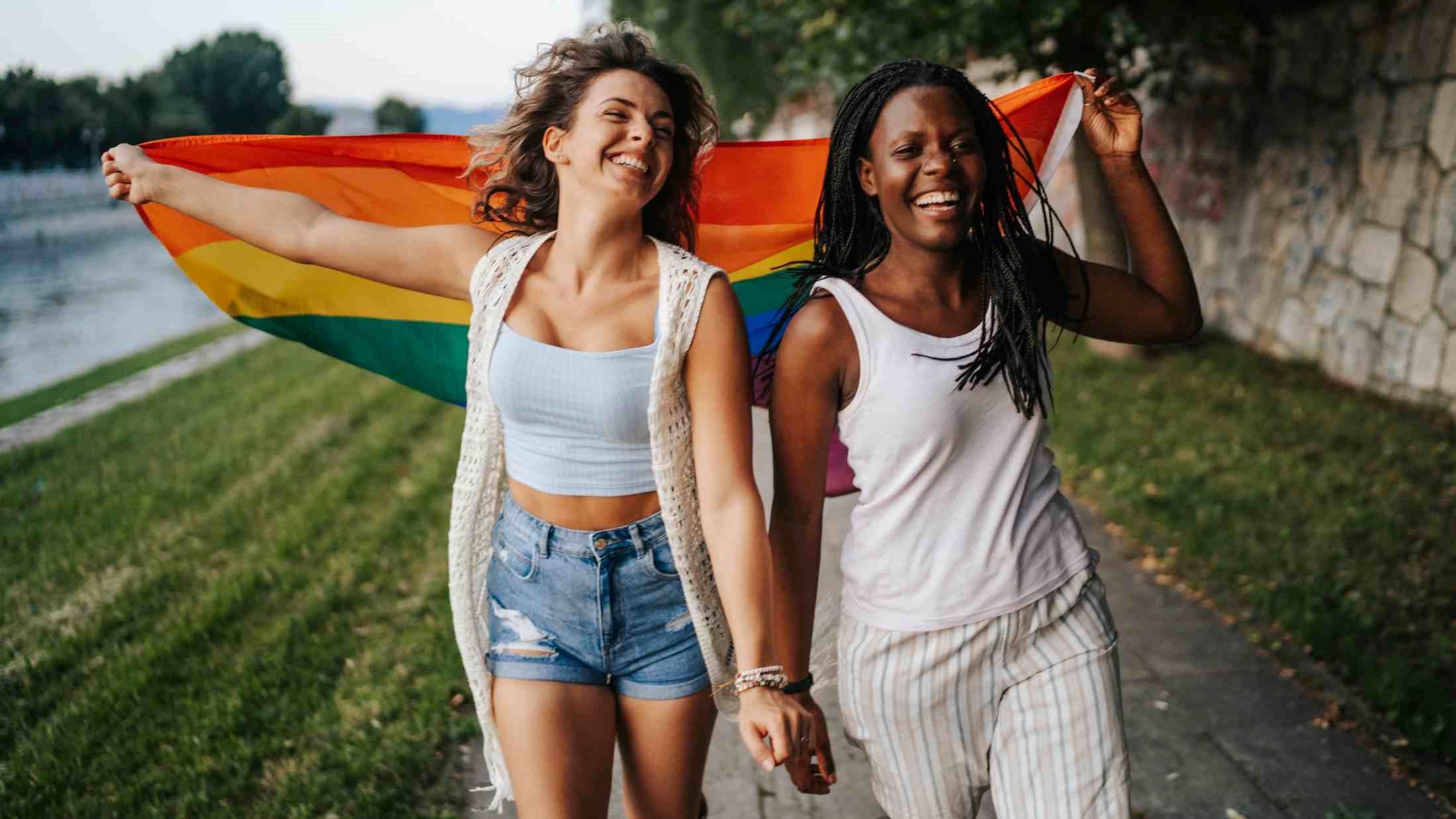 Young multi ethnic lesbian couple with rainbow flag running on city quay
